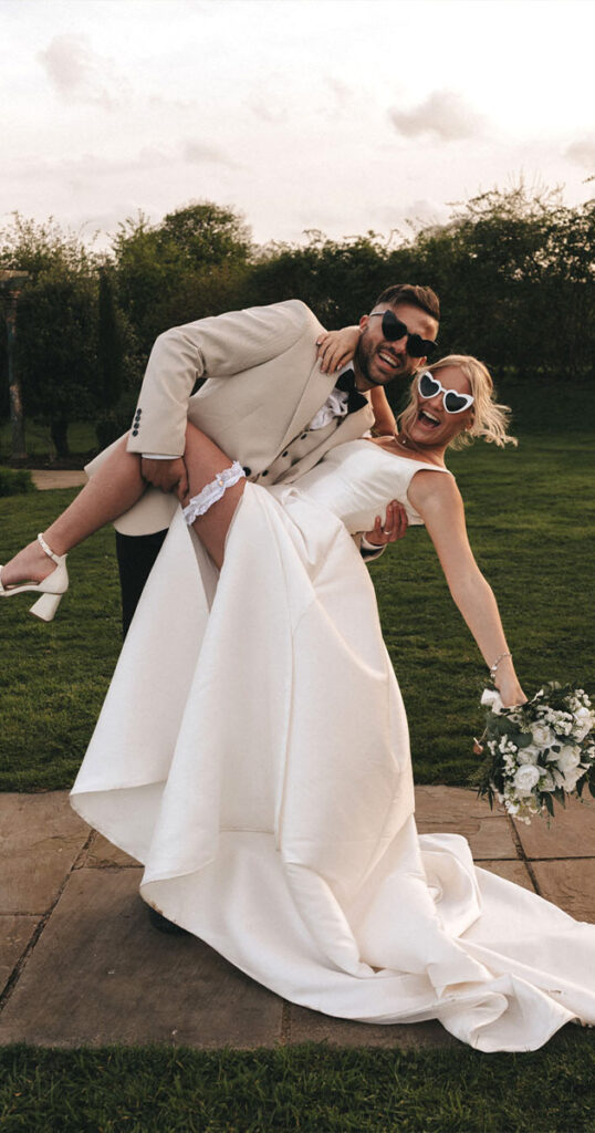 A joyful couple poses outdoors, capturing a perfect photo moment at their Brackenborough Hotel wedding. The groom, in a beige suit and sunglasses, playfully lifts the bride in her white gown and heart-shaped sunglasses. She holds a bouquet and shows her garter on a charming stone path surrounded by grass and trees. © Aimee Lince Photography