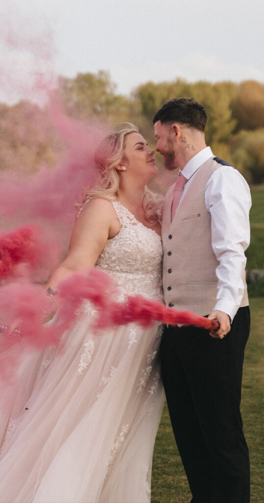 A bride and groom stand closely on a grassy field at Kenwick Park, each holding pink smoke flares that create a vibrant cloud around them. The bride wears a lace wedding gown, and the groom is in a vest and tie. They gaze affectionately at each other, with trees blurred in the background. © Aimee Lince Photography