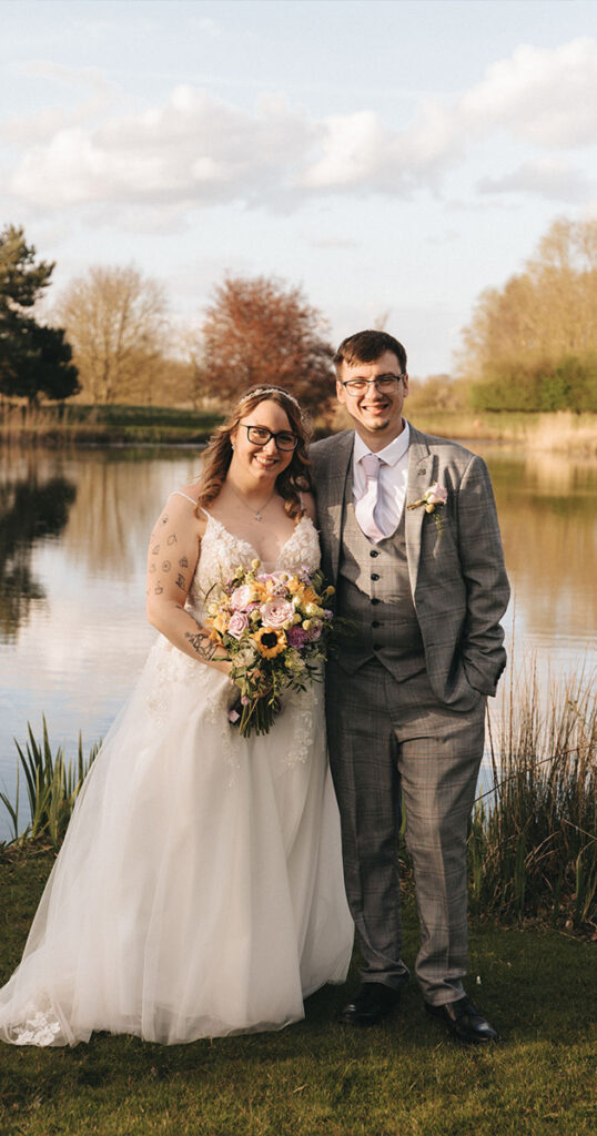 A bride and groom stand smiling by a serene lake at Hall Farm Hotel. The bride wears a white gown and holds a bouquet of sunflowers and pastel flowers, while the groom is in a gray suit with a light vest and tie. Surrounded by greenery, this moment captures the essence of wedding photography amid nature's beauty. © Aimee Lince Photography