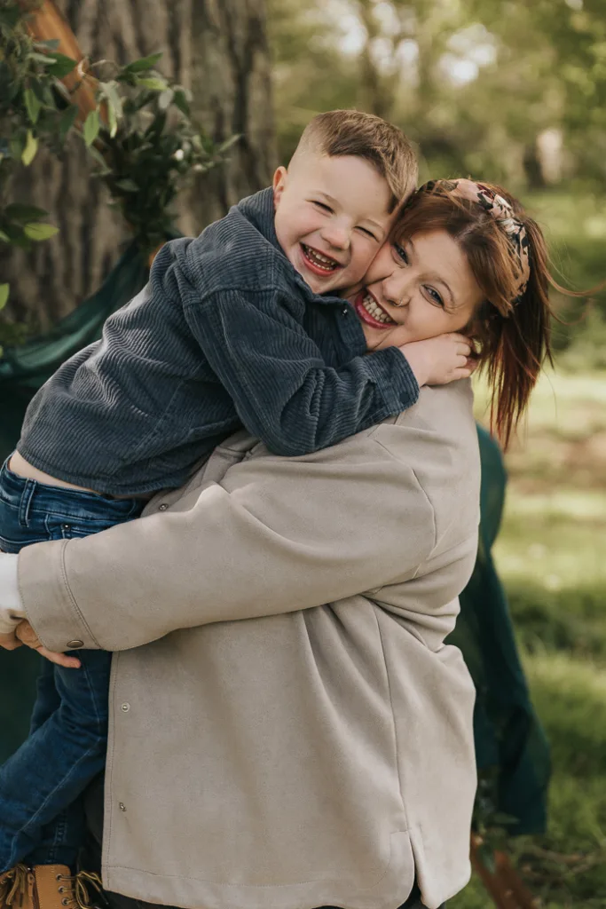 A smiling woman in a light coat holds a young boy with Down syndrome, both laughing joyfully under a tree in Weelsby Woods. Dressed in a blue jacket and jeans, the child revels in the family photos mini session on this bright, sunny day as leaves and branches blur into the background. © Aimee Lince Photography - Wedding photographer in Lincolnshire, Yorkshire & Nottinghamshire