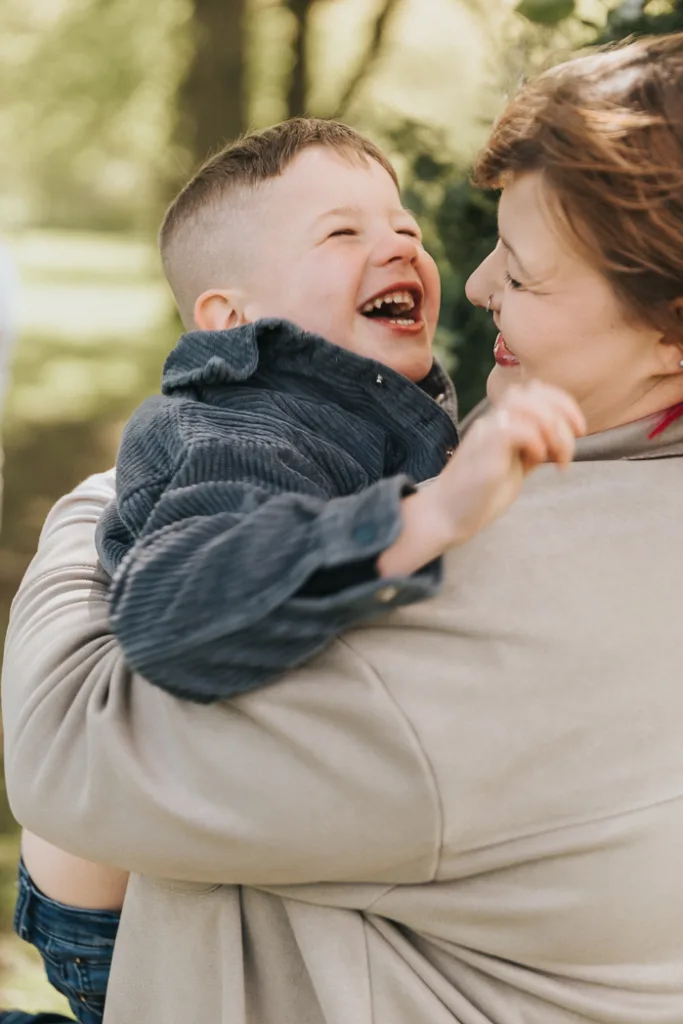 A woman holds a laughing young boy with short hair in her arms amidst the lush greenery of Weelsby Woods. The child wears a dark jacket, and the woman is in a light-colored coat. Both smile brightly, sharing a joyful moment during their sunny family photo mini session. © Aimee Lince Photography - Wedding photographer in Lincolnshire, Yorkshire & Nottinghamshire
