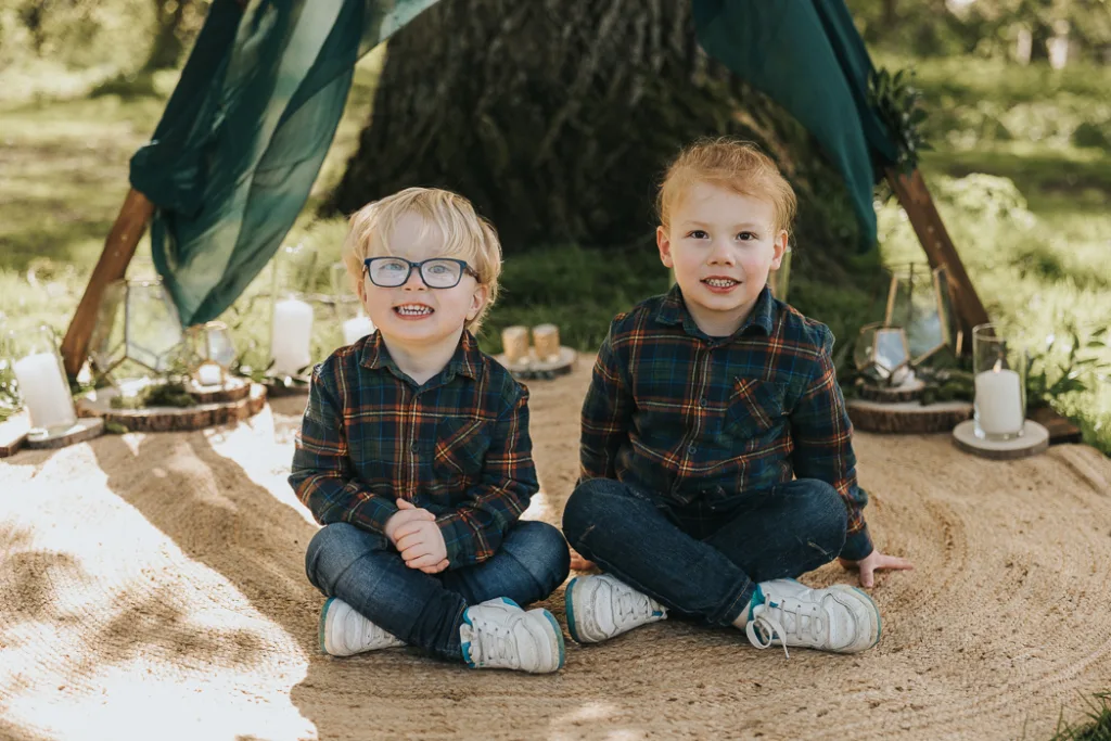 Two young children sit cross-legged on a woven rug in Weelsby Woods, smiling in their matching plaid shirts and jeans. Behind them stands a makeshift tent with green fabric, surrounded by candles on wooden bases. Sunlight filters through the trees, casting dappled shadows during this enchanting mini family session. © Aimee Lince Photography - Wedding photographer in Lincolnshire, Yorkshire & Nottinghamshire