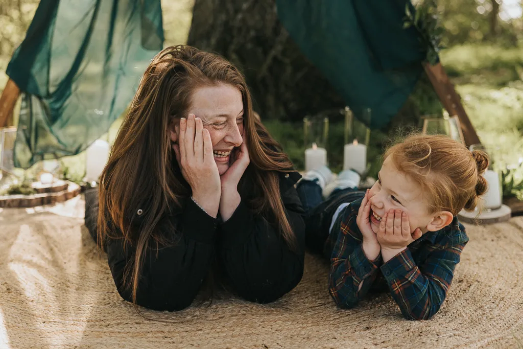 A woman and child lie on a woven mat, propped on their elbows, sharing smiles. Their cheeks rest on hands as the sun filters through the trees. In Weelsby Woods, with candles in glass holders and sheer fabric draped nearby, it's a perfect setting for enchanting mini family photo sessions. © Aimee Lince Photography - Wedding photographer in Lincolnshire, Yorkshire & Nottinghamshire