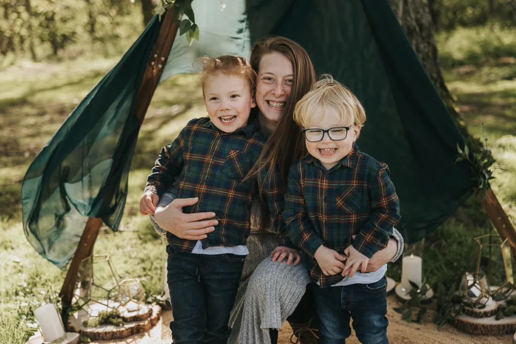 A smiling woman kneels outdoors in front of a dark green tent with two laughing children, both in matching plaid shirts and jeans. Sunlight filters through the trees, illuminating the joyful group—perfect for cherished family photos. © Aimee Lince Photography - Wedding photographer in Lincolnshire, Yorkshire & Nottinghamshire