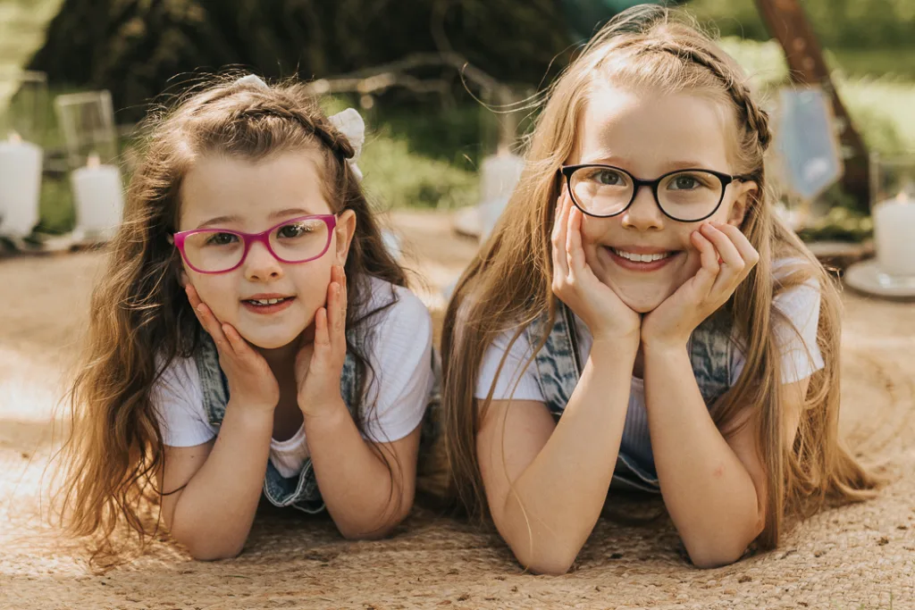 Two young girls with long hair, both wearing glasses, lie on their stomachs on a woven mat in Weelsby Woods. Dressed in white shirts and denim overalls, they smile with hands under chins. Lit candles in glass holders glow behind them against the backdrop of lush green grass—perfect for family photos. © Aimee Lince Photography - Wedding photographer in Lincolnshire, Yorkshire & Nottinghamshire