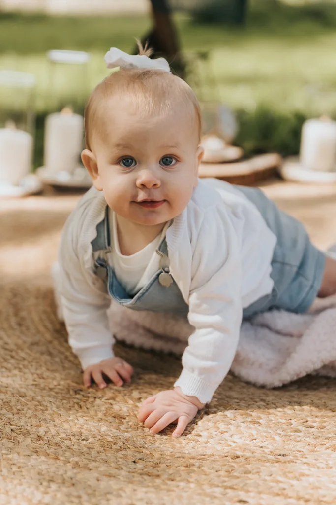 A baby with blue eyes and light hair is crawling on a textured woven rug in Weelsby Woods. They wear a light blue overall, a white sweater, and a small white bow. The background is blurred with candles and greenery, perfect for intimate family photos in this serene outdoor setting. © Aimee Lince Photography - Wedding photographer in Lincolnshire, Yorkshire & Nottinghamshire