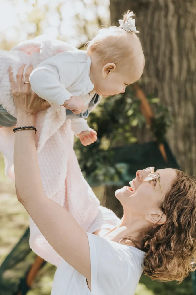 A joyful woman with curly hair lifts a smiling baby above her in Weelsby Woods. The baby, wrapped in a pink blanket, wears a white outfit and bow. The woman sports sunglasses and a white shirt as trees and greenery softly blur the sunny background—a perfect moment for family photos. © Aimee Lince Photography - Wedding photographer in Lincolnshire, Yorkshire & Nottinghamshire