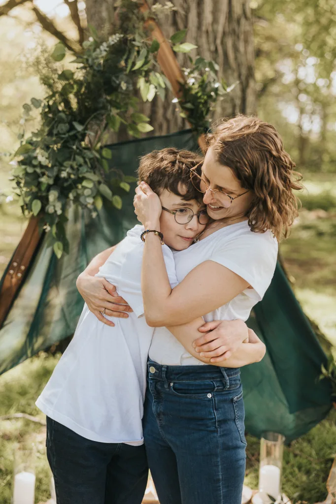 In the tranquil setting of Weelsby Woods, a young boy with glasses and a woman with wavy hair share a warm embrace. Both in white tops and jeans, they stand among leafy branches and a green cloth backdrop. The scene is peaceful and joyful, perfect for family photos. © Aimee Lince Photography - Wedding photographer in Lincolnshire, Yorkshire & Nottinghamshire
