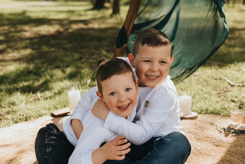 Two young boys in white long-sleeve shirts hug and smile while sitting on a blanket outdoors during a family photos mini session. Behind them, a small tent with draped fabric stands in Weelsby Woods' grassy clearing, where soft sunlight filters through trees and candles flicker gently. © Aimee Lince Photography - Wedding photographer in Lincolnshire, Yorkshire & Nottinghamshire