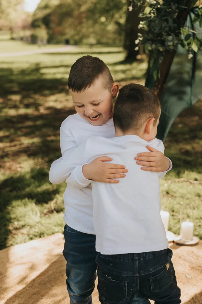 In a warm and playful scene at Weelsby Woods, two young boys joyfully hug on a grassy area. Clad in white long-sleeve shirts and dark pants, one boy faces the camera with laughter while the other turns his back. Sunlight filters through the trees, capturing a perfect family photo moment. © Aimee Lince Photography - Wedding photographer in Lincolnshire, Yorkshire & Nottinghamshire