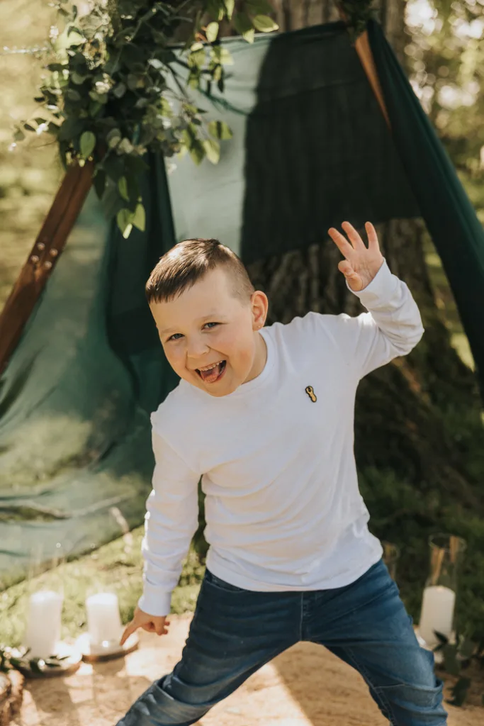 A young boy with a short haircut, wearing a white long-sleeve shirt and jeans, playfully poses with a big smile and one hand raised. He stands in front of a green tent at Weelsby Woods, surrounded by grass and trees, with candles placed on the ground near him—perfect for mini family photo sessions. © Aimee Lince Photography - Wedding photographer in Lincolnshire, Yorkshire & Nottinghamshire