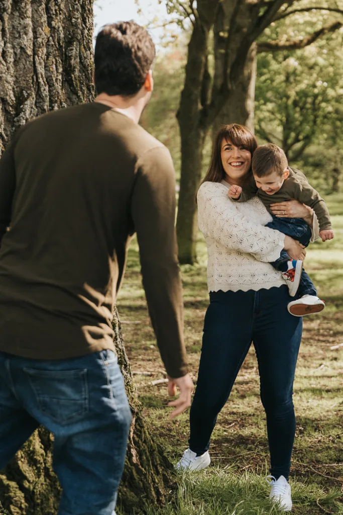 A woman in a white sweater holds a smiling child wearing a green jacket, both facing a man in a green shirt by a tree. They're outdoors in Weelsby Woods amidst green grass and trees, capturing playful family bonding perfect for cherished family photos. © Aimee Lince Photography - Wedding photographer in Lincolnshire, Yorkshire & Nottinghamshire