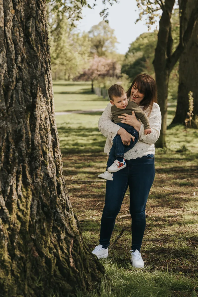 A woman in a white sweater and jeans holds a smiling young boy, both dressed casually, as they pose for family photos near a large tree. The sunlit park, reminiscent of Weelsby Woods, with green grass and scattered shadows, sets the scene for a peaceful and joyful mini session outdoors. © Aimee Lince Photography - Wedding photographer in Lincolnshire, Yorkshire & Nottinghamshire