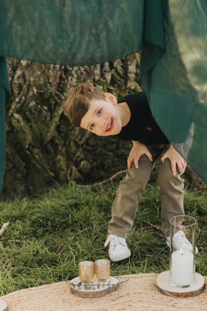 A young boy with short brown hair, wearing a black sweater and khaki pants, is playfully peeking under a green canopy at Weelsby Woods. He's smiling with his head tilted. Below him on the grass is a round beige mat with two gold candle holders and a tall glass vase, perfect for family photos. © Aimee Lince Photography - Wedding photographer in Lincolnshire, Yorkshire & Nottinghamshire