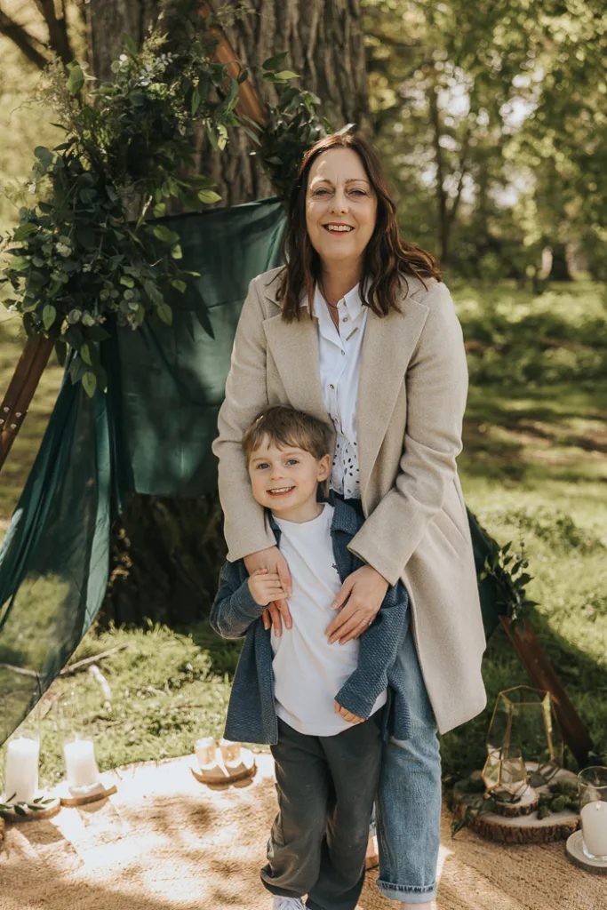 A woman in a beige coat smiles while hugging a young boy, clad in a blue jacket and white shirt. They stand in Weelsby Woods, surrounded by the lush canopy of trees. Behind them, a dark green fabric is draped with leafy decorations as candles and small lanterns illuminate this enchanting family photo setting. © Aimee Lince Photography - Wedding photographer in Lincolnshire, Yorkshire & Nottinghamshire
