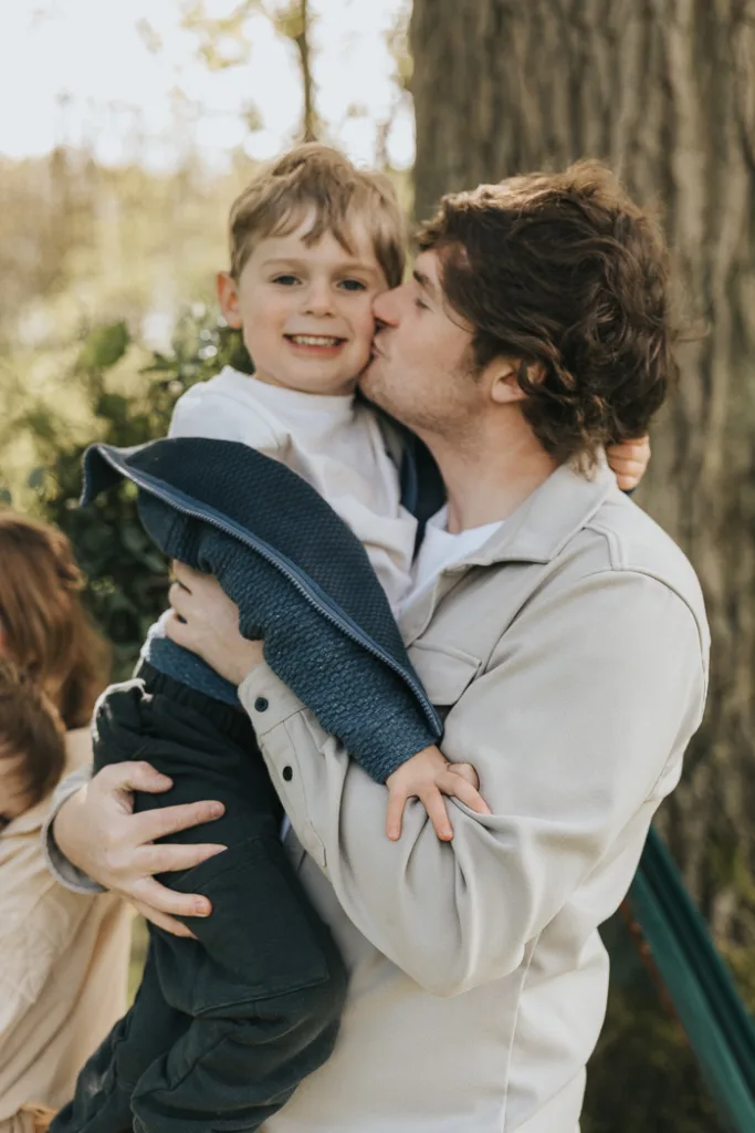 A man affectionately kisses the cheek of a smiling child he's holding. The man is wearing a light jacket, and the child is wrapped in a blue knit blanket. They're enjoying family photos in Weelsby Woods, with a large tree trunk and greenery creating a warm, cheerful atmosphere. © Aimee Lince Photography - Wedding photographer in Lincolnshire, Yorkshire & Nottinghamshire