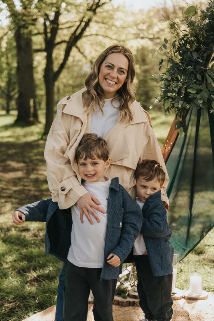 A woman smiles standing outdoors with two young boys during mini sessions at Weelsby Woods. She wears a tan coat and white shirt, while the boys sport blue jackets. They're on a picnic blanket amid greenery and a decorative arch, with sunlight filtering through the trees, creating a warm, natural setting perfect for family photos. © Aimee Lince Photography - Wedding photographer in Lincolnshire, Yorkshire & Nottinghamshire