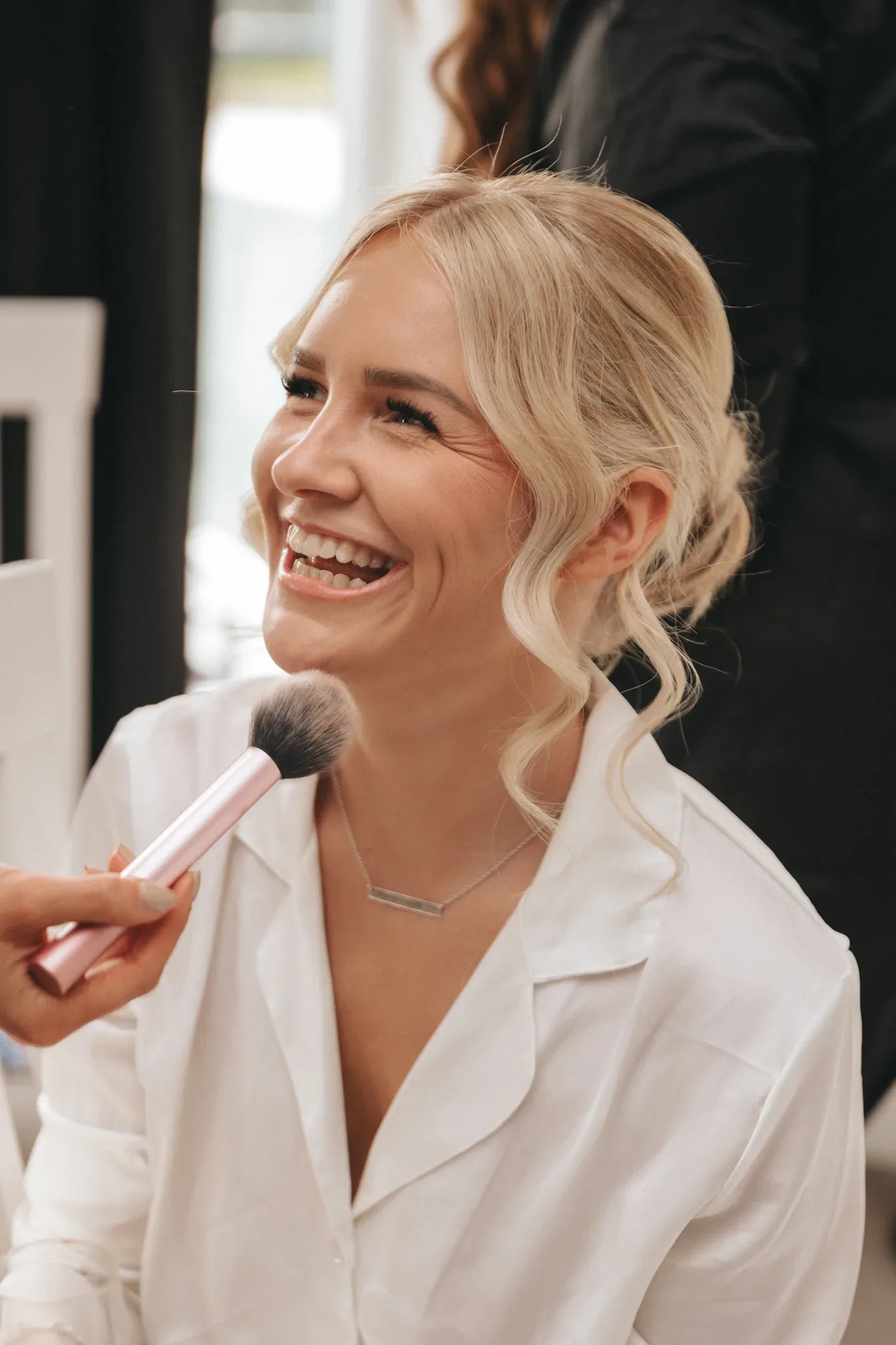 A joyful woman having makeup applied, with curly blond hair and a white shirt, laughing while a makeup artist uses a brush on her cheek in a brightly lit room.