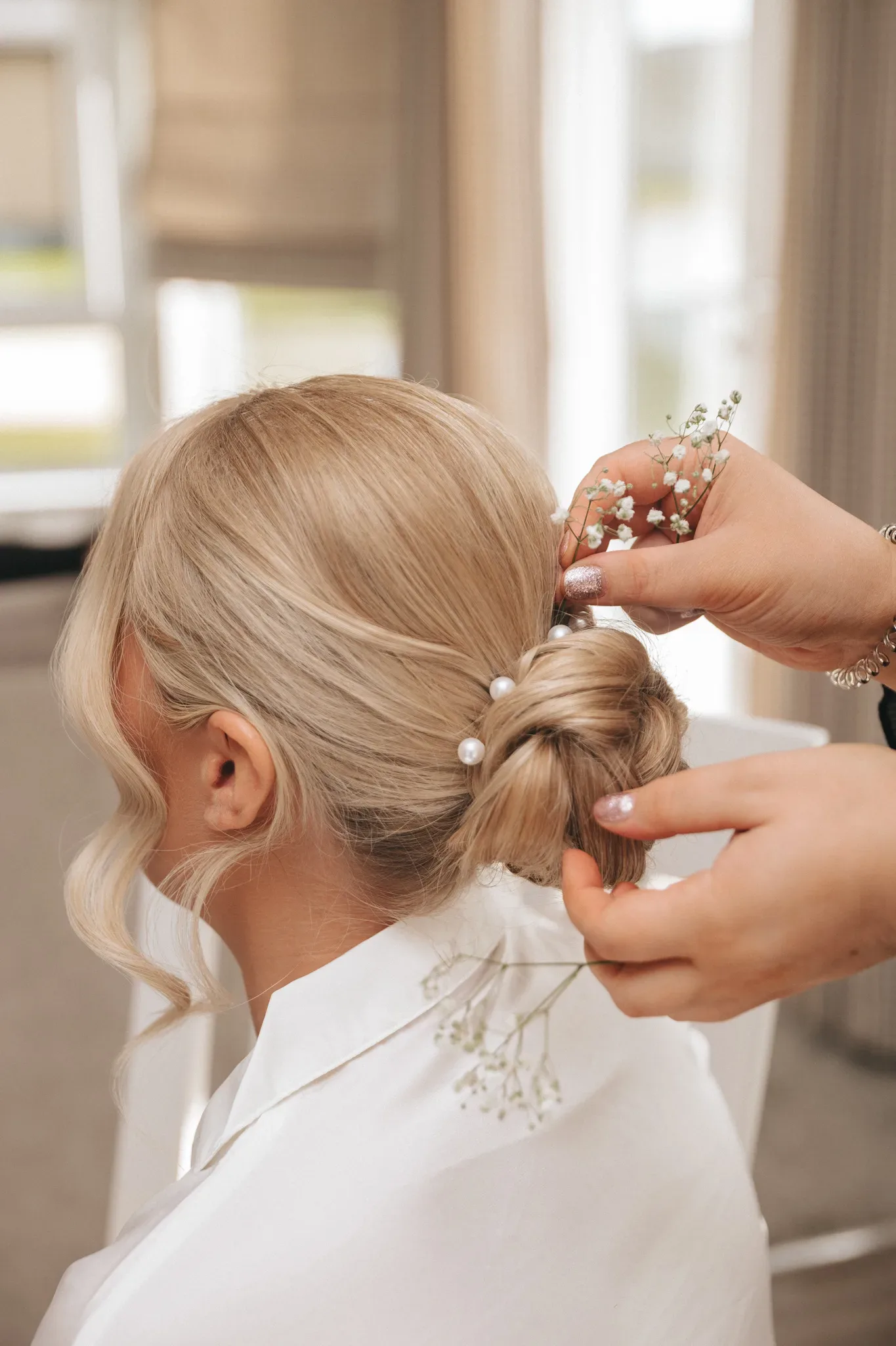A hairstylist arranges a blonde woman's hair into an elegant updo, adding small white flowers and pearls. the setting suggests a bright, airy room.