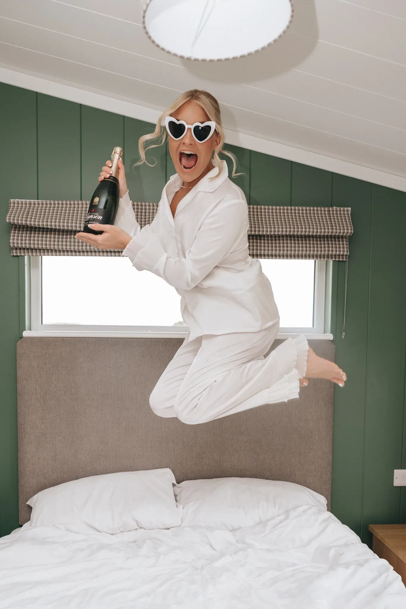 A joyful woman in white pajamas and heart-shaped sunglasses jumps on a bed, holding a champagne bottle. her mouth is open in mid-cheer, and the room has a cozy, green-walled interior with a window above the headboard.