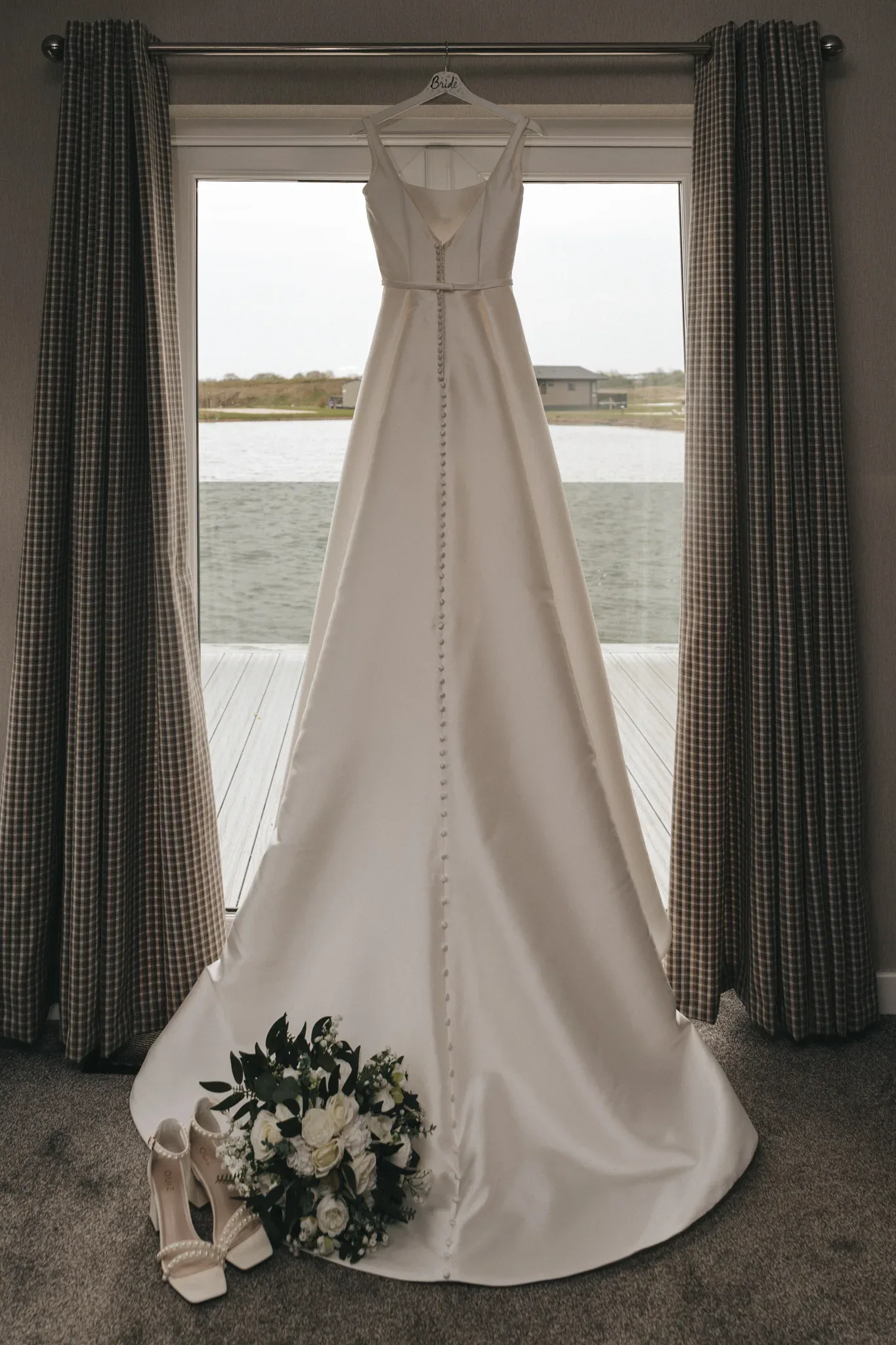A wedding dress hangs in front of a window framed by curtains, overlooking a lake. below, a pair of white bridal shoes and a bouquet of white flowers with green leaves are placed on the carpet.