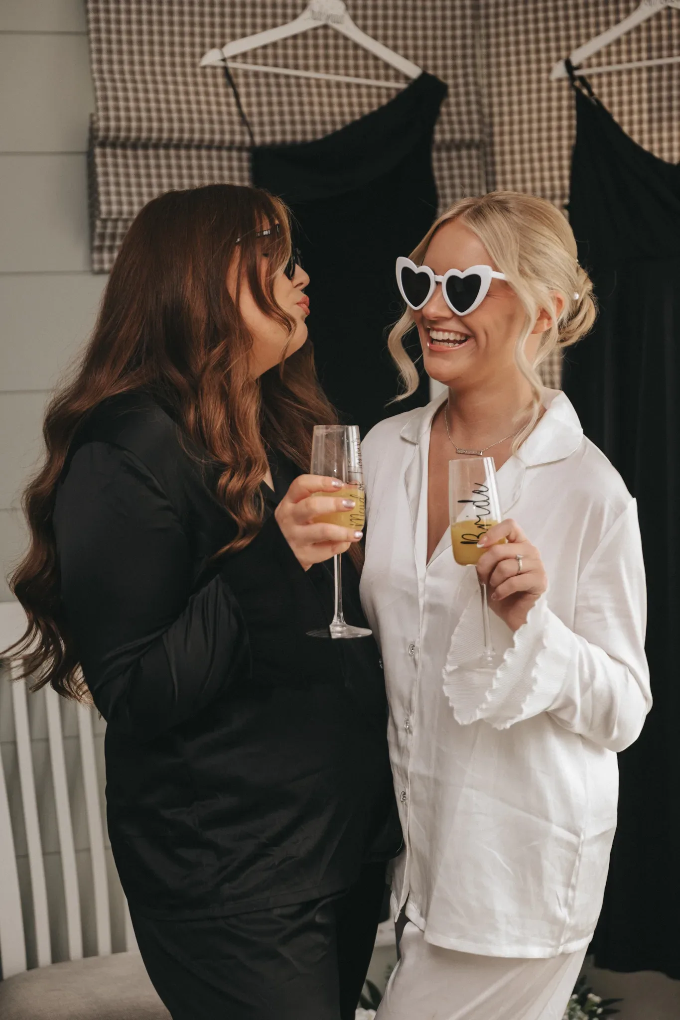 Two women, one with heart-shaped sunglasses, smiling and clinking champagne glasses, dressed in stylish black and white outfits, standing in a room with a patterned backdrop.