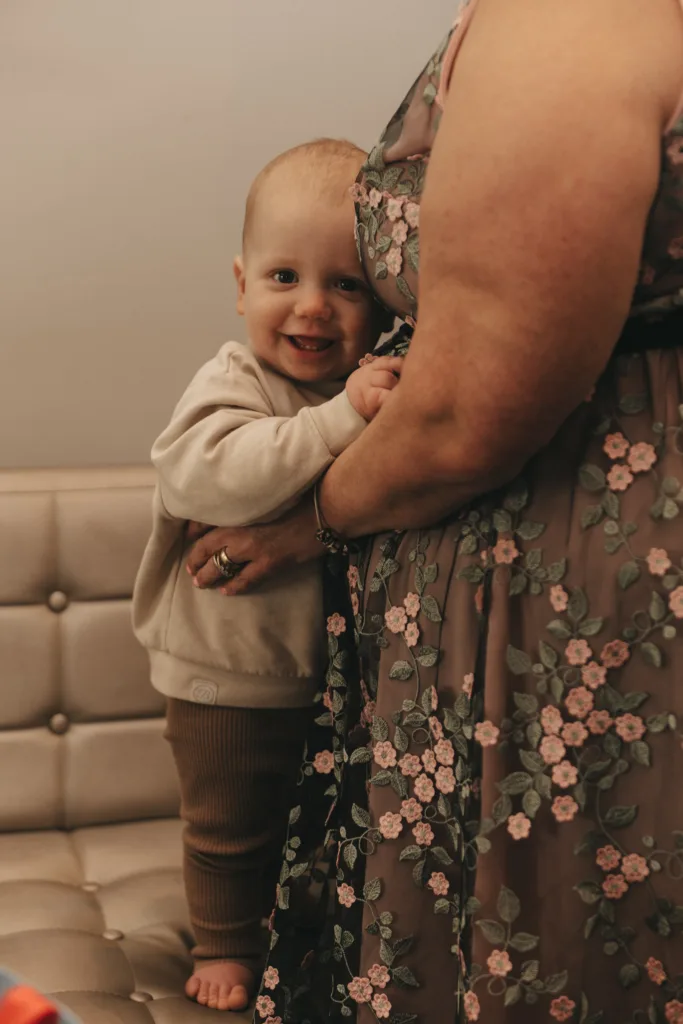 A baby with a joyful smile clings to an adult, standing next to a tufted sofa. the baby wears a light hoodie and brown pants; the adult is in a floral dress.