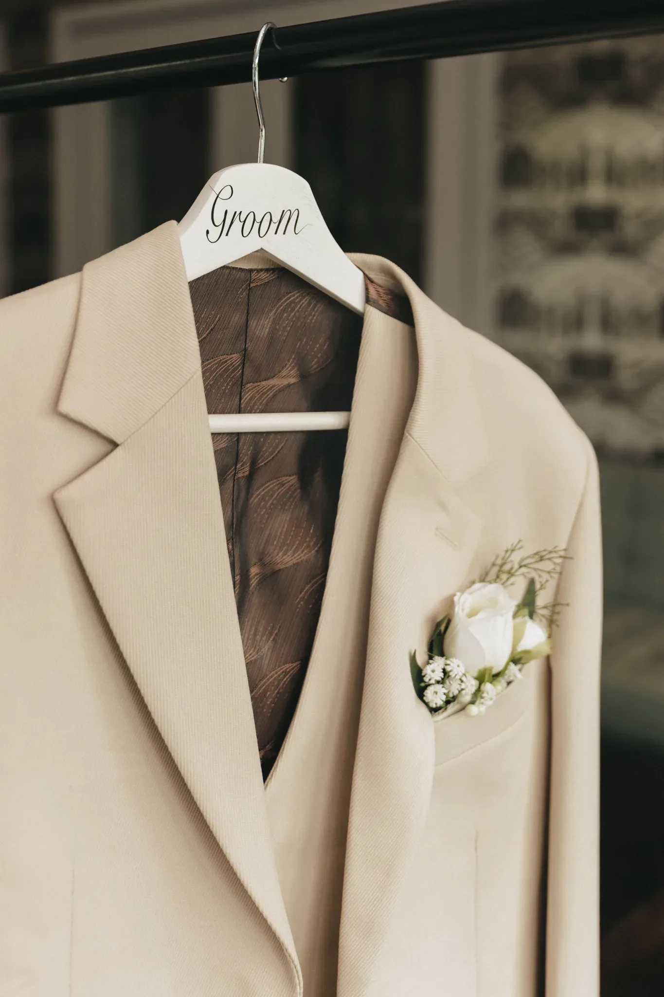 A beige groom's suit hangs on a wooden hanger labeled "groom," featuring a boutonniere with white roses and greenery on the lapel, set against a softly blurred background.