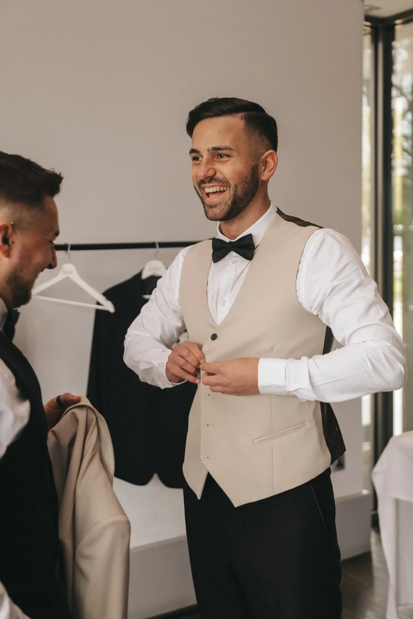 A joyful groom in a beige vest and white shirt smiles broadly, adjusting his black bow tie, with his reflection visible in a mirror held by another man in the background. the setting suggests a well-lit room with a modern design.