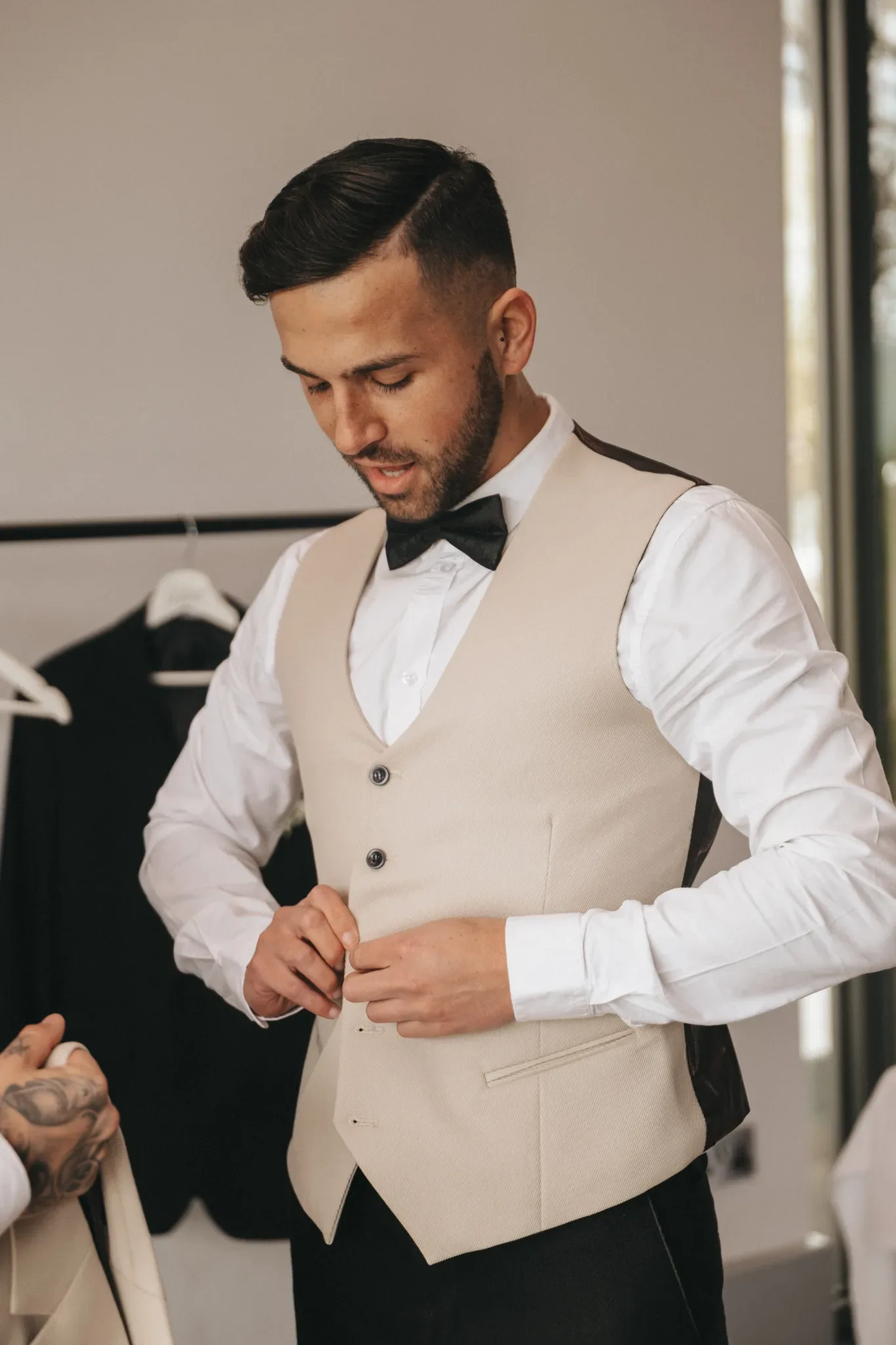 A groom in a white dress shirt and beige vest buttons his vest, preparing for his wedding. he has short, styled hair and a well-groomed beard, and is focused on his task.