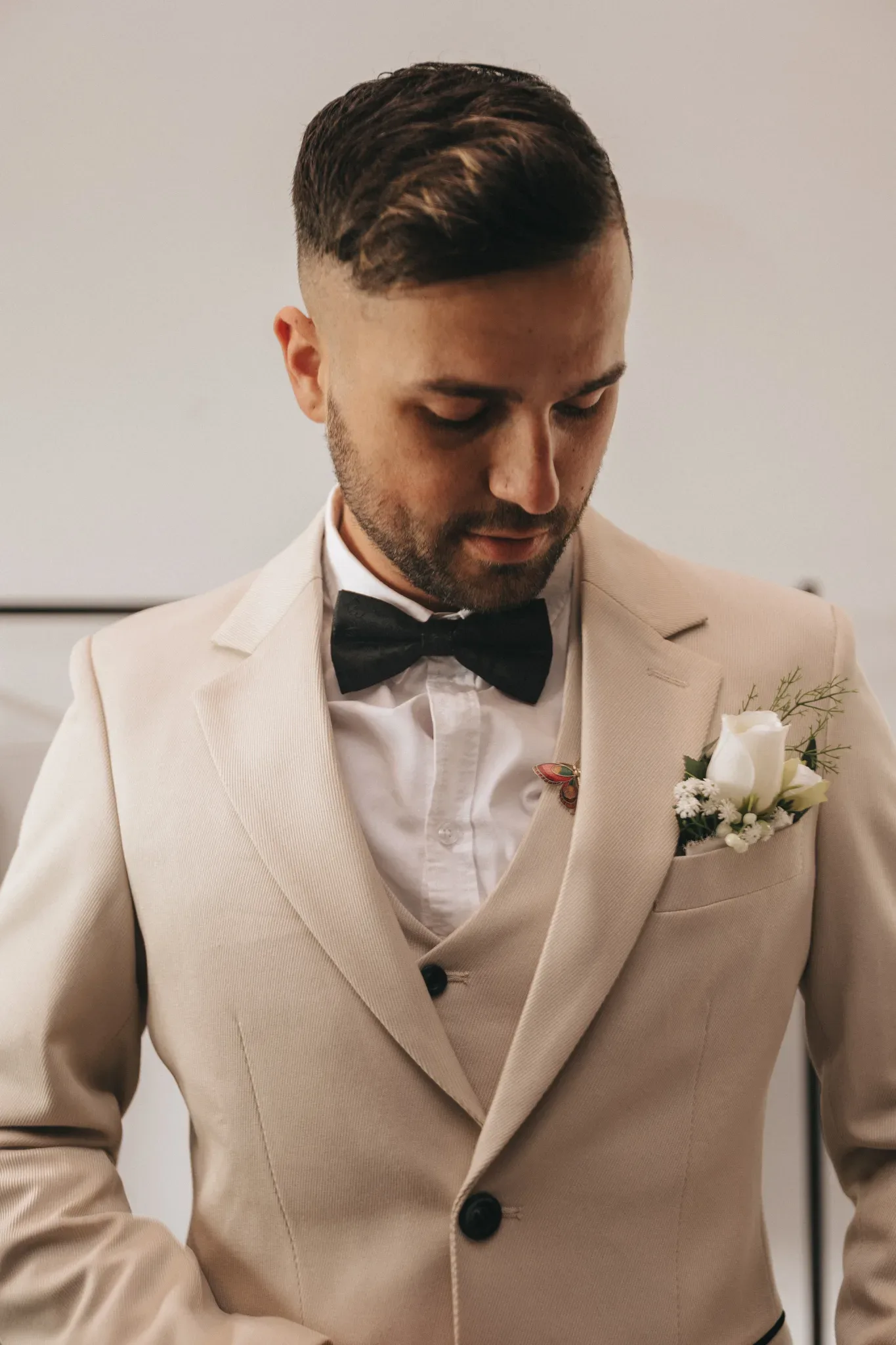 A man in a beige suit with a black bow tie and boutonniere looks downward, his expression thoughtful. close up captures elegant details of his stylish haircut and formal attire.