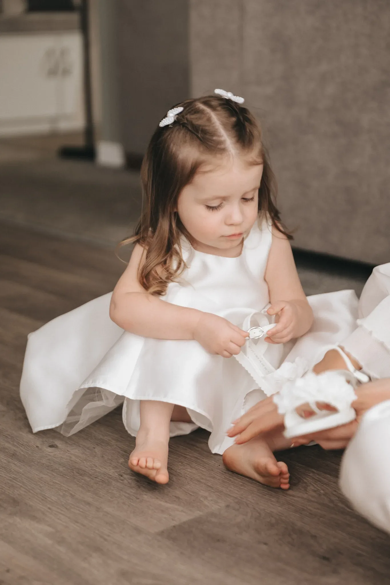 A young girl in a white satin dress with delicate hair bands sits on a wooden floor, intently fastening a white ribbon on a woman's shoe, who is cropped at the knees.