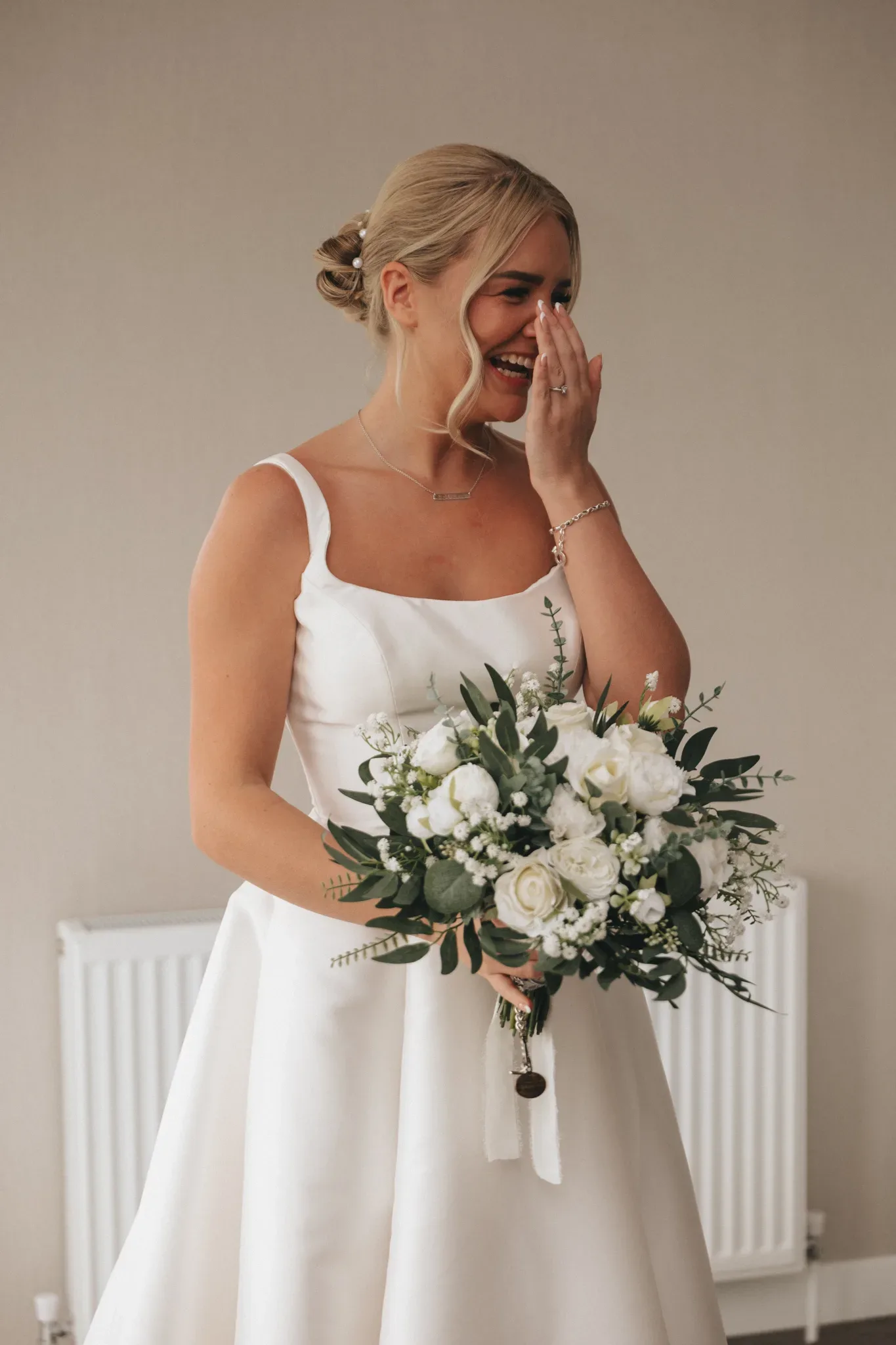 A bride in a white dress with spaghetti straps holds a bouquet of white flowers and greenery, looking emotional and wiping tears from her face, standing indoors with a neutral background.