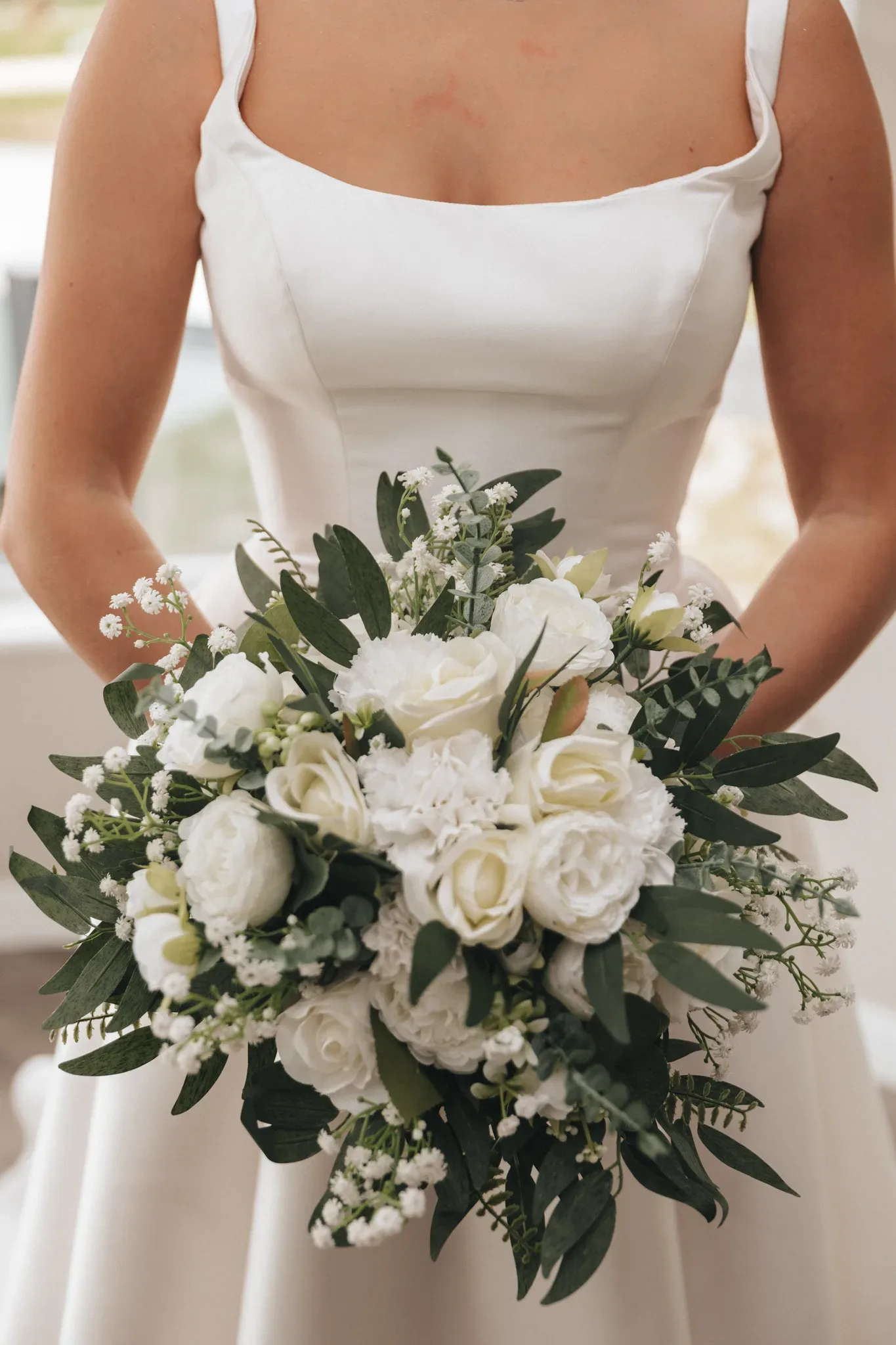 A bride in a white sleeveless dress holds a large bouquet of white roses and green foliage, showcasing the elegant simplicity of her attire and the delicate floral arrangement.