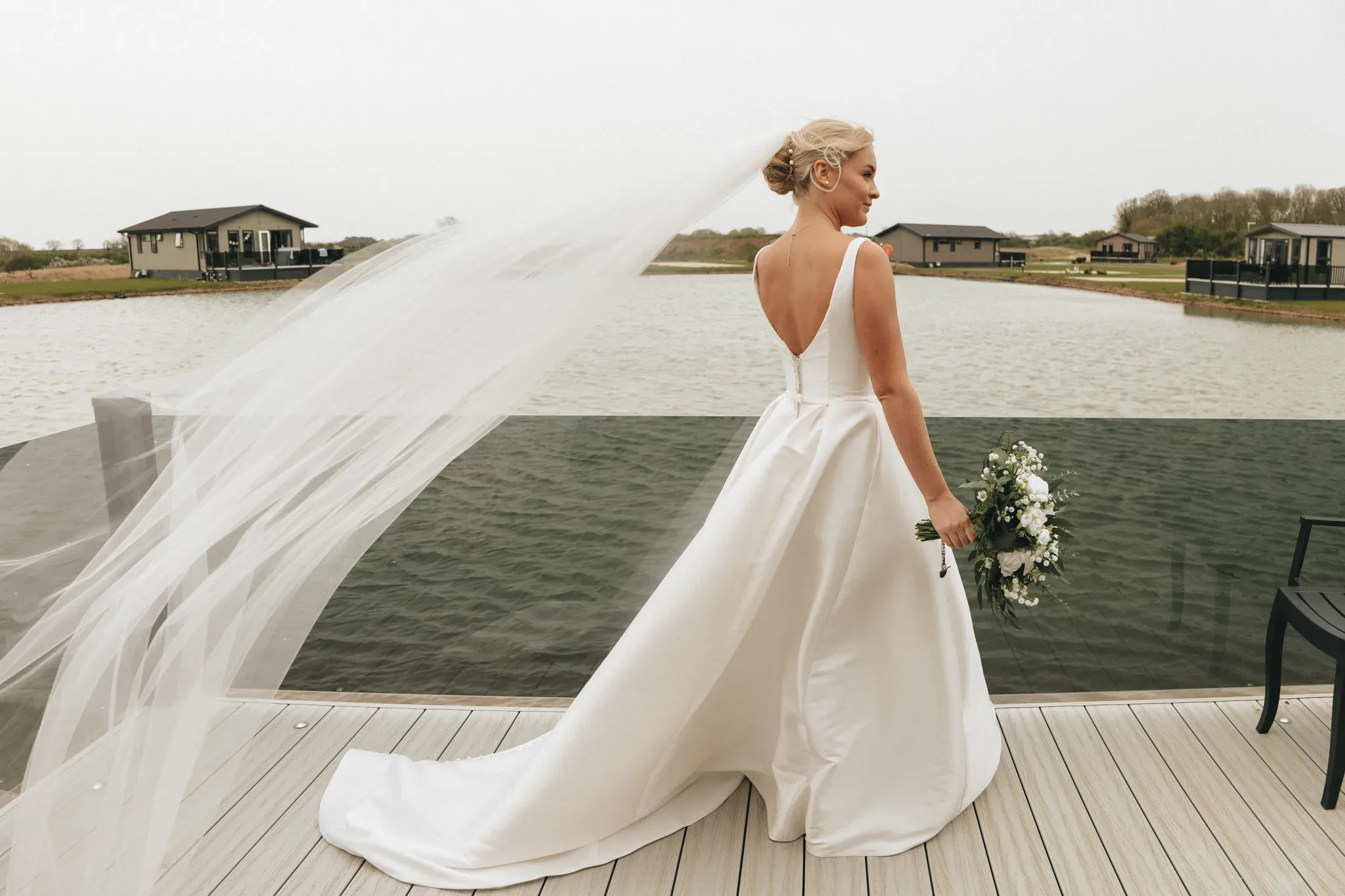 A bride in a white dress with a flowing veil stands on a lakeside deck, holding a bouquet. her back is to the camera, showcasing the elegant design of her dress against a serene lake and cloudy sky backdrop.