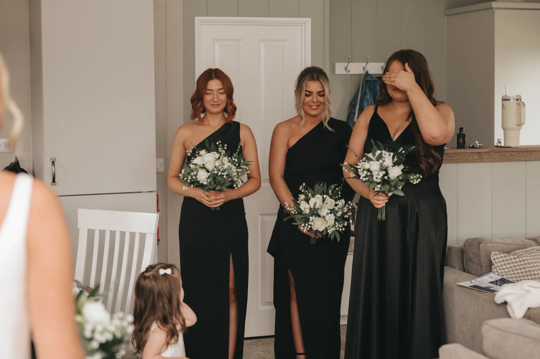 Three bridesmaids in black dresses hold bouquets, standing in a room with modern decor. the bridesmaid on the right covers her face, possibly emotional, while the others smile gently.