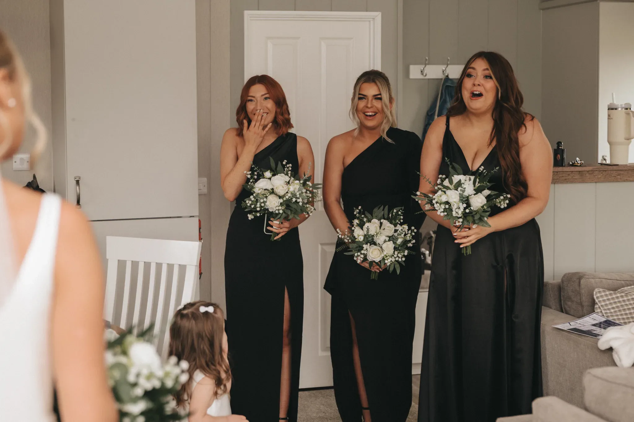 Three women in elegant black dresses hold white bouquets and express joy and surprise, watching a bride in the foreground in a home setting.