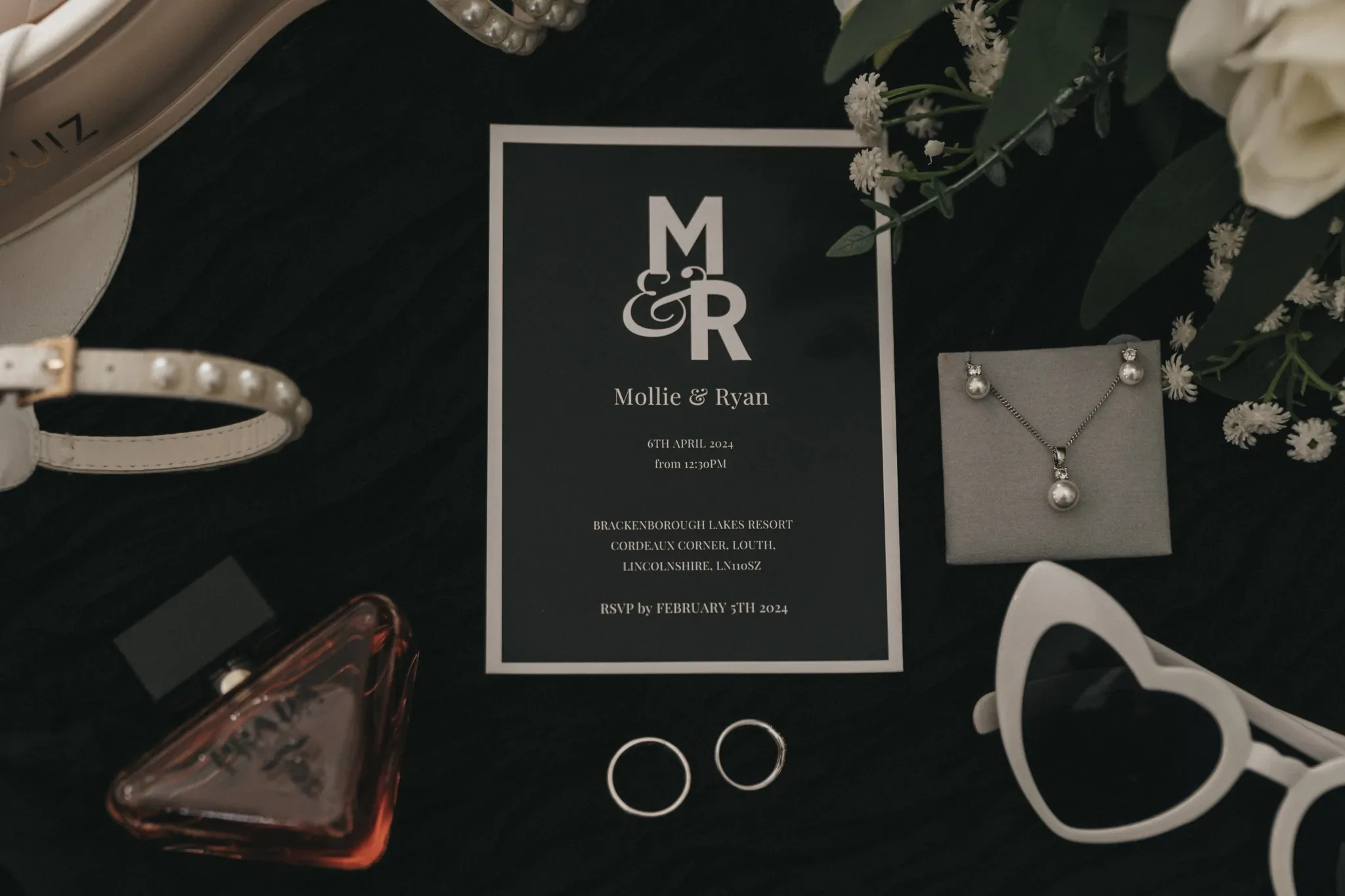Elegant wedding invitation on a black background with "mollie & ryan" in large letters, surrounded by floral decorations, a white strap with pearls, a pair of earrings, sunglasses, and a perfume bottle.