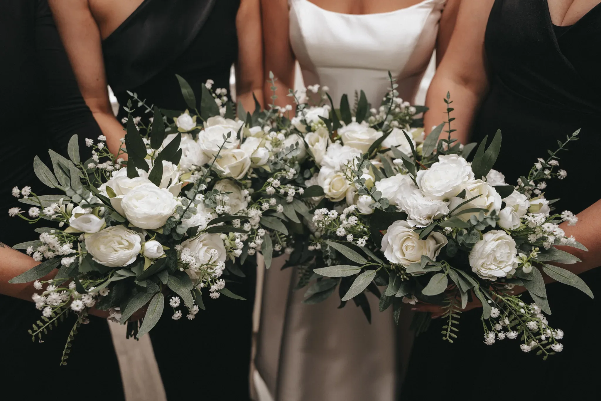 Three women in elegant white and black dresses holding large bouquets with white roses and lush greenery close to the camera, focusing on the flowers.