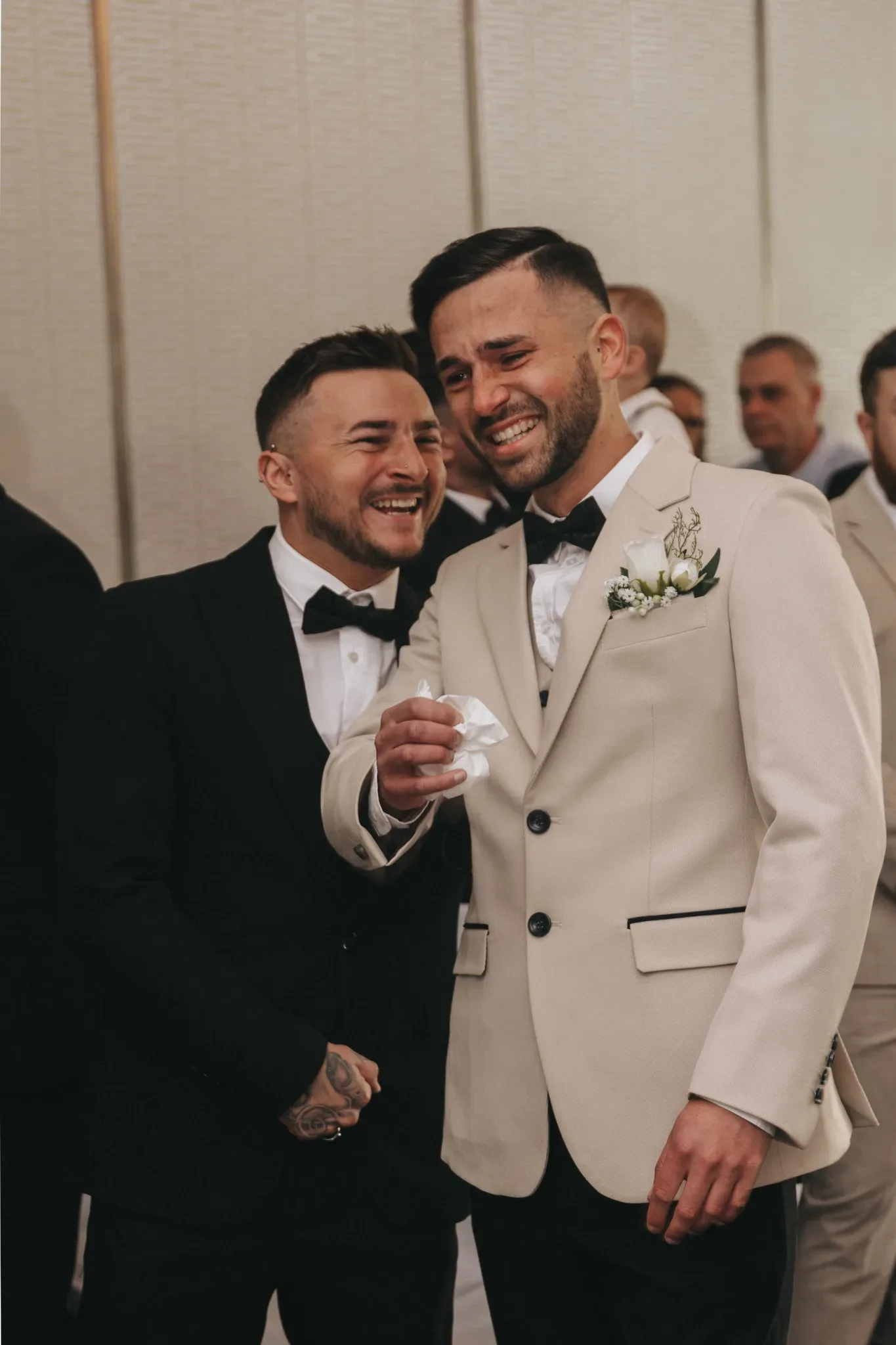 Two men in formal wedding attire, one in a black tuxedo and the other in a cream suit, smile joyfully at a wedding. the man in black wipes away a tear, hinting at an emotional moment.