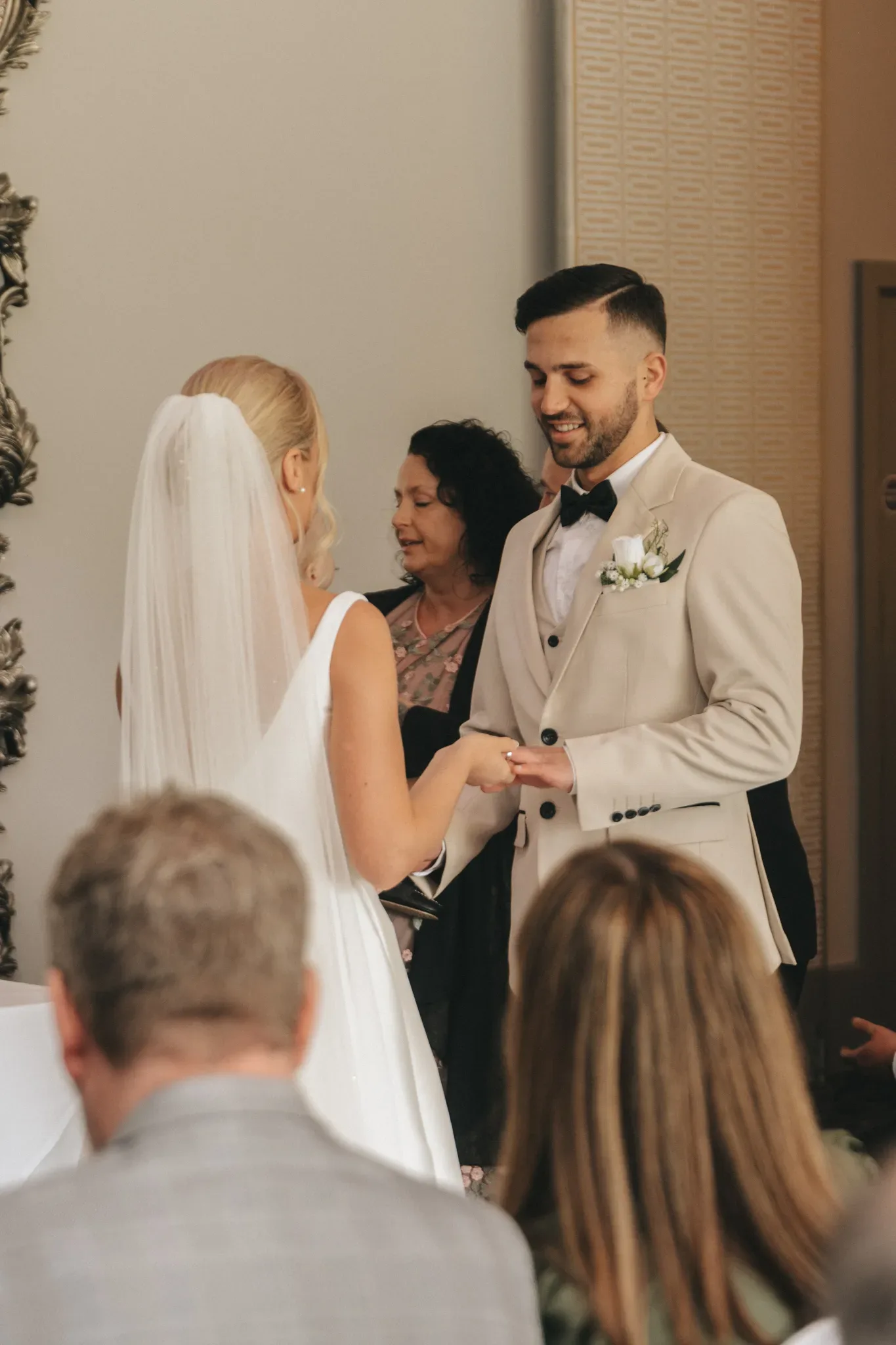 A bride and groom hold hands and exchange rings during their wedding ceremony, with an officiant smiling behind them. the groom is in a beige suit, while the bride wears a white dress and veil.