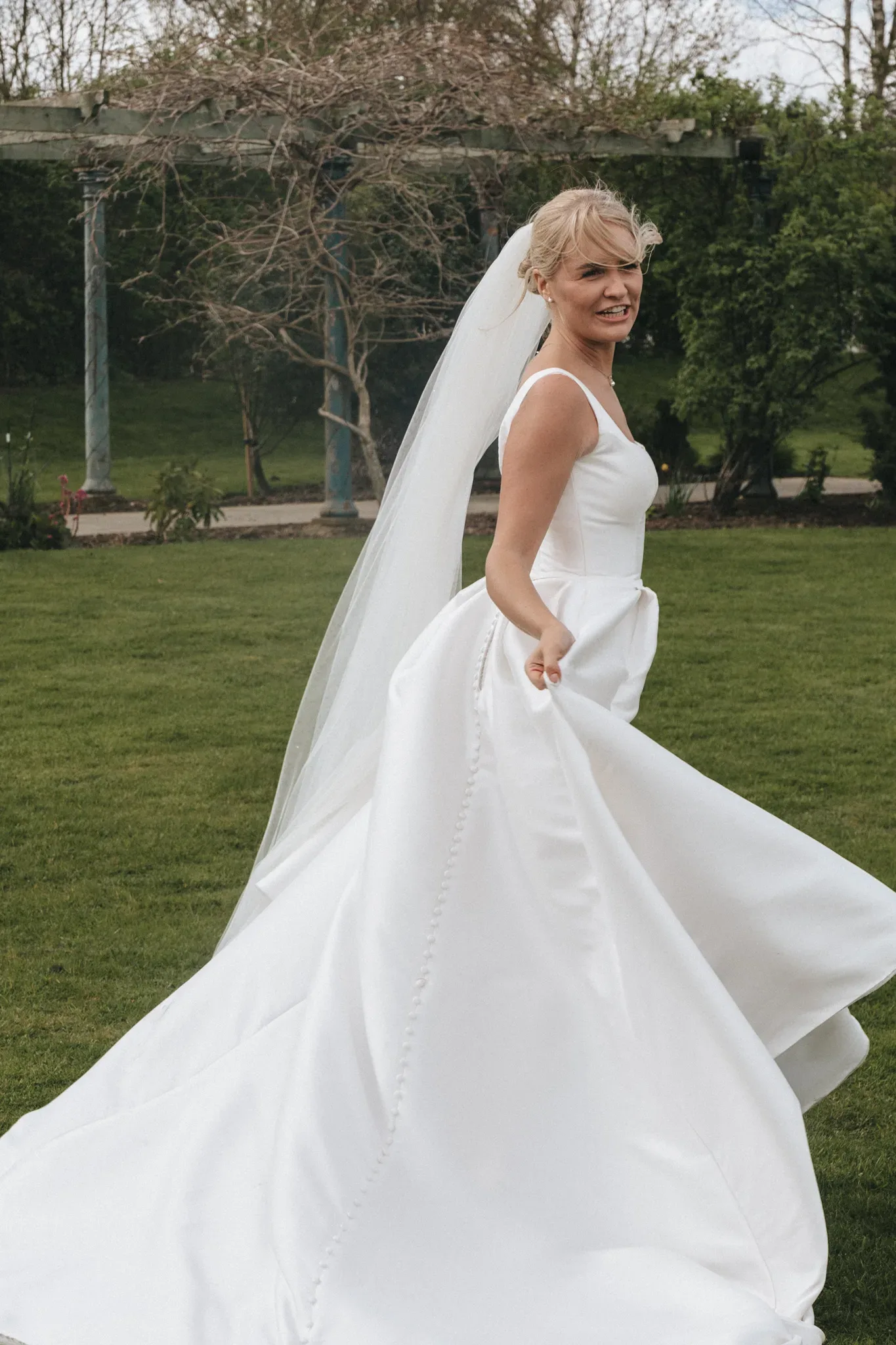 A bride in a white wedding dress with a flowing veil smiles over her shoulder while walking in a lush garden, her dress billowing gently behind her.