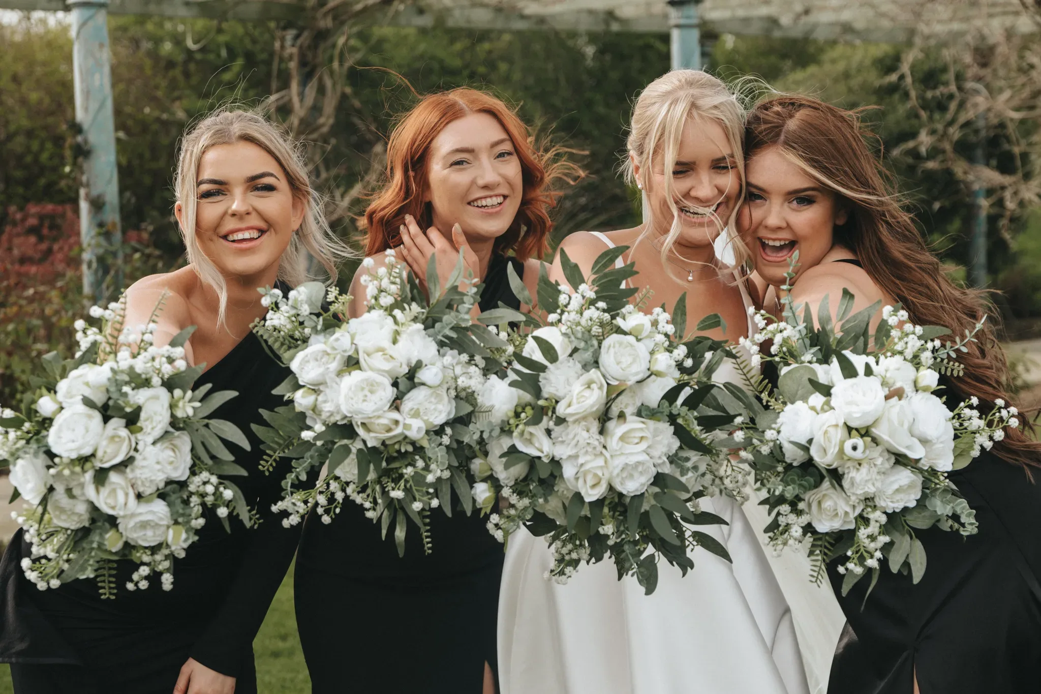 Four smiling women in formal wear, holding bouquets with white flowers, posing together at an outdoor event, likely a wedding. the bride in the center wears white, while the others wear black.
