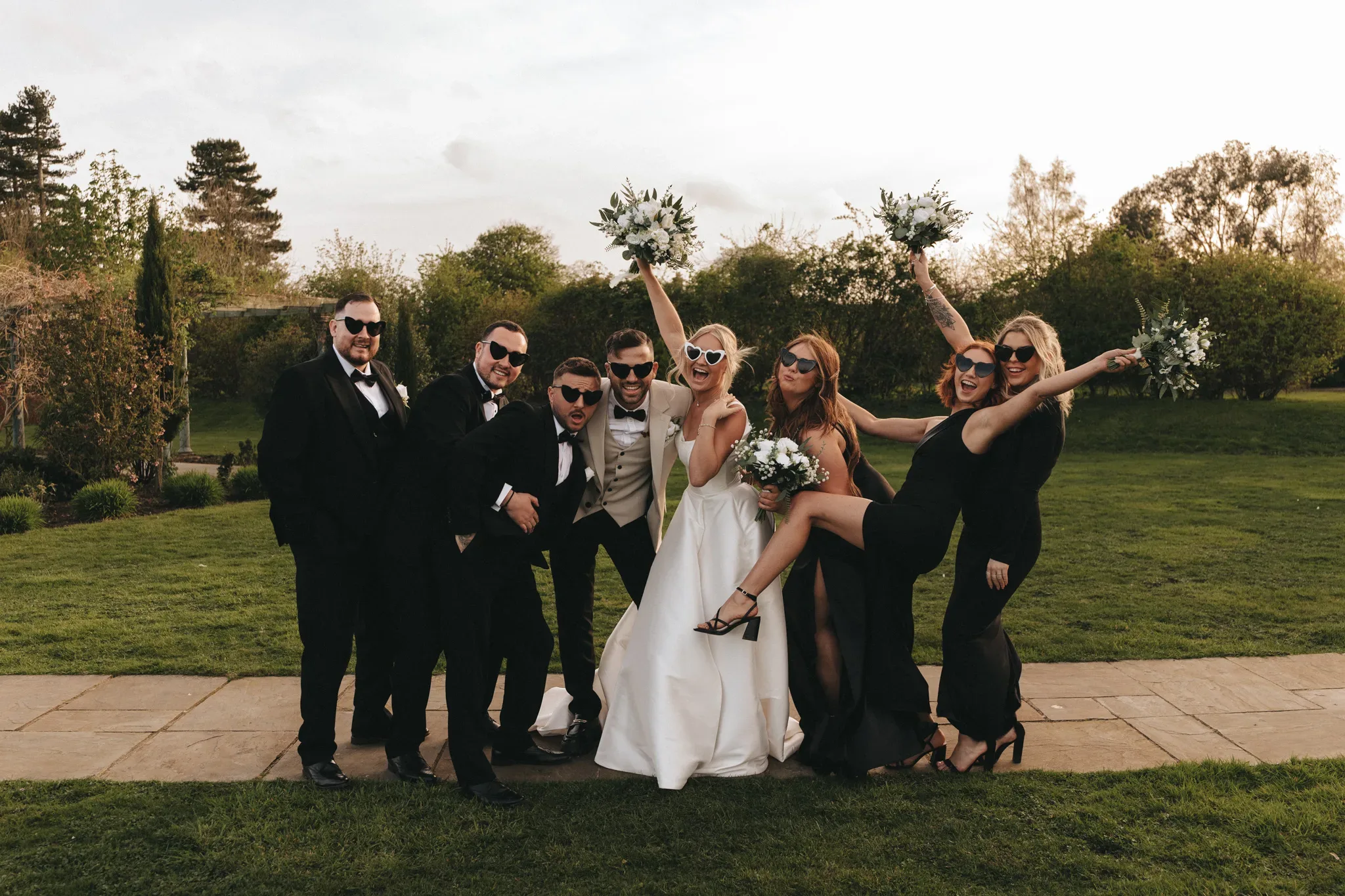 A joyful wedding party outside, featuring a bride and groom in the center surrounded by four bridesmaids and three groomsmen, all dressed elegantly in black, celebrating with playful poses on a grassy field at sunset. Aimee Lince, Kind Words.