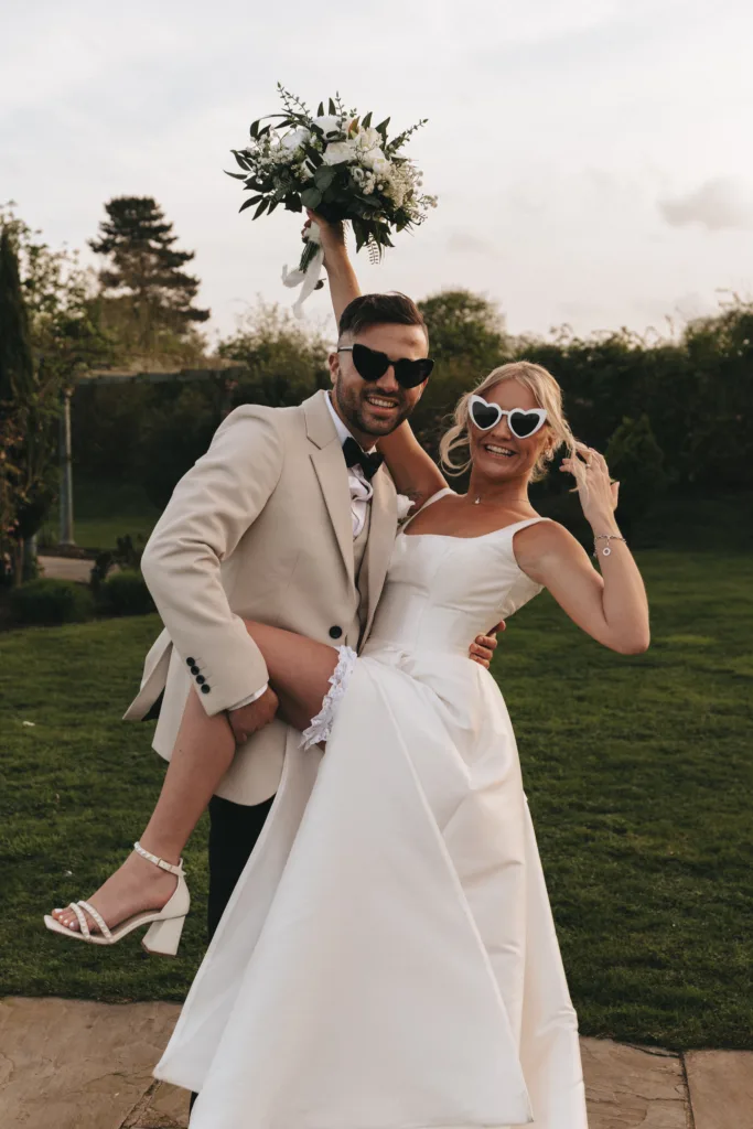 A joyful bride and groom pose outdoors in formal wedding attire. the groom, lifting the bride, wears a beige suit and sunglasses, while the bride in a white dress and matching sunglasses holds a bouquet aloft.