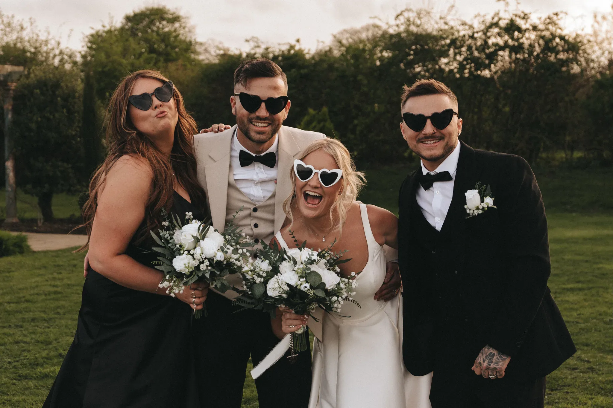 Two couples in formal wedding attire pose joyfully outdoors, each wearing playful sunglasses. the men wear black tuxedos and the woman in a white dress holds a bouquet, with greenery in the background.