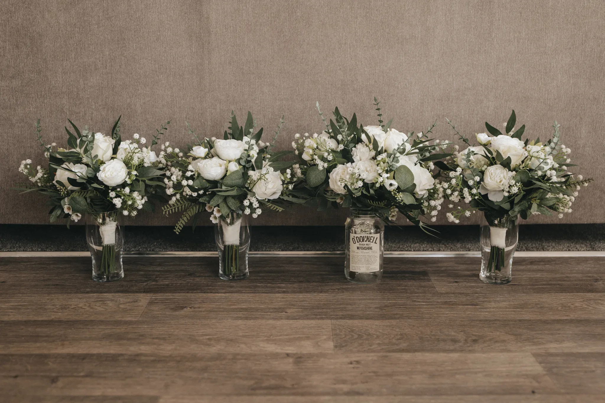 Four bouquets in glass jars, featuring white flowers and greenery, are placed on a wooden floor against a gray textured wall. one jar in the center has a "gordon's" gin label.