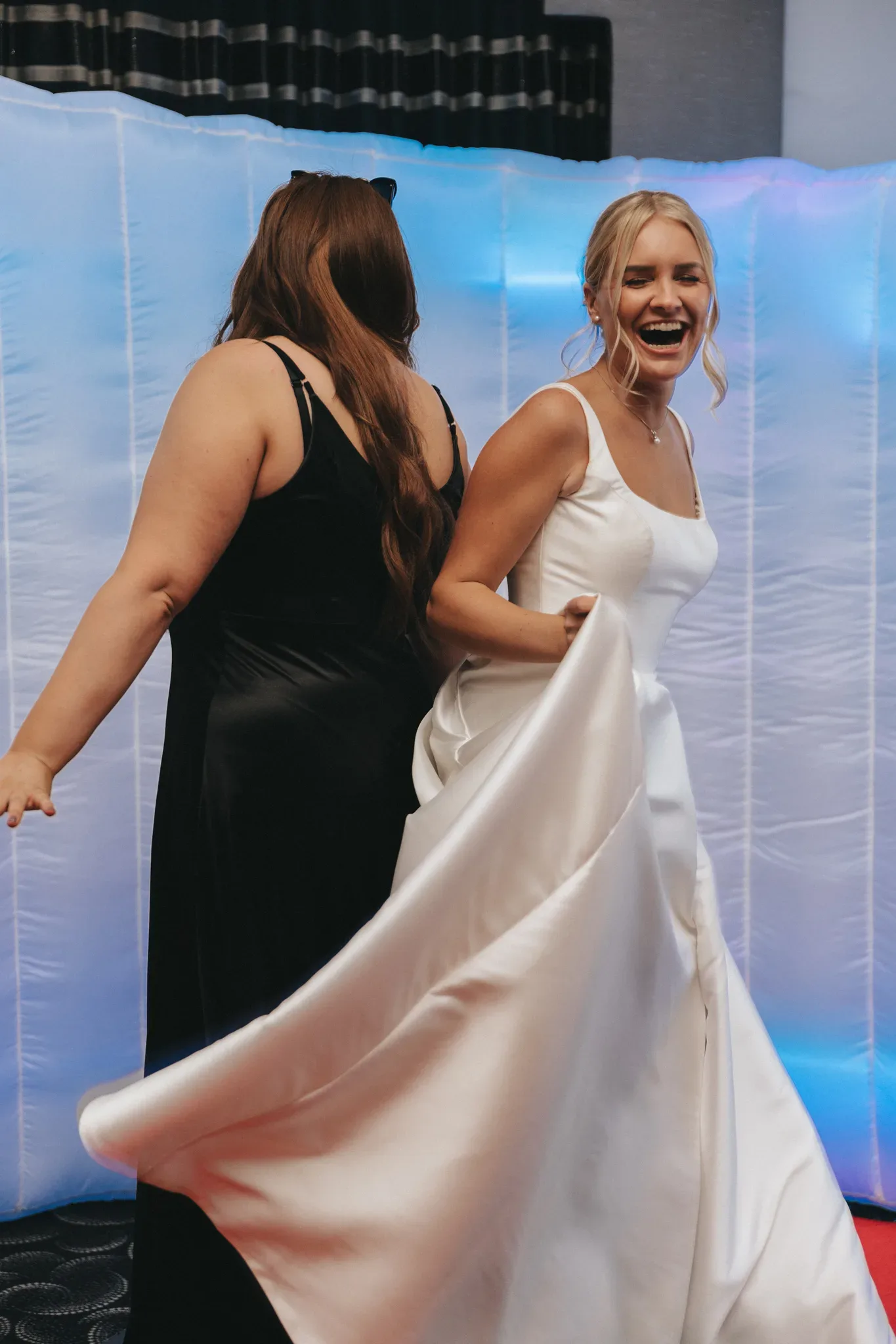 Two women dancing joyfully at an event. the woman on the right, dressed in a white gown, laughs while holding her dress, and the woman on the left, in a black dress, dances beside her.