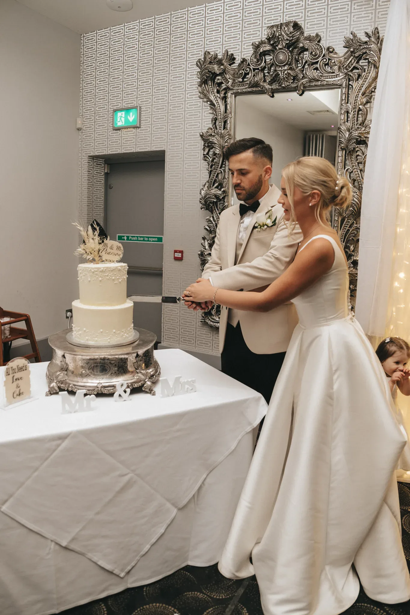 A bride and groom are cutting their wedding cake together in a decorated room. there is an ornate silver frame in the background and a child peeking out from behind the table on the right.