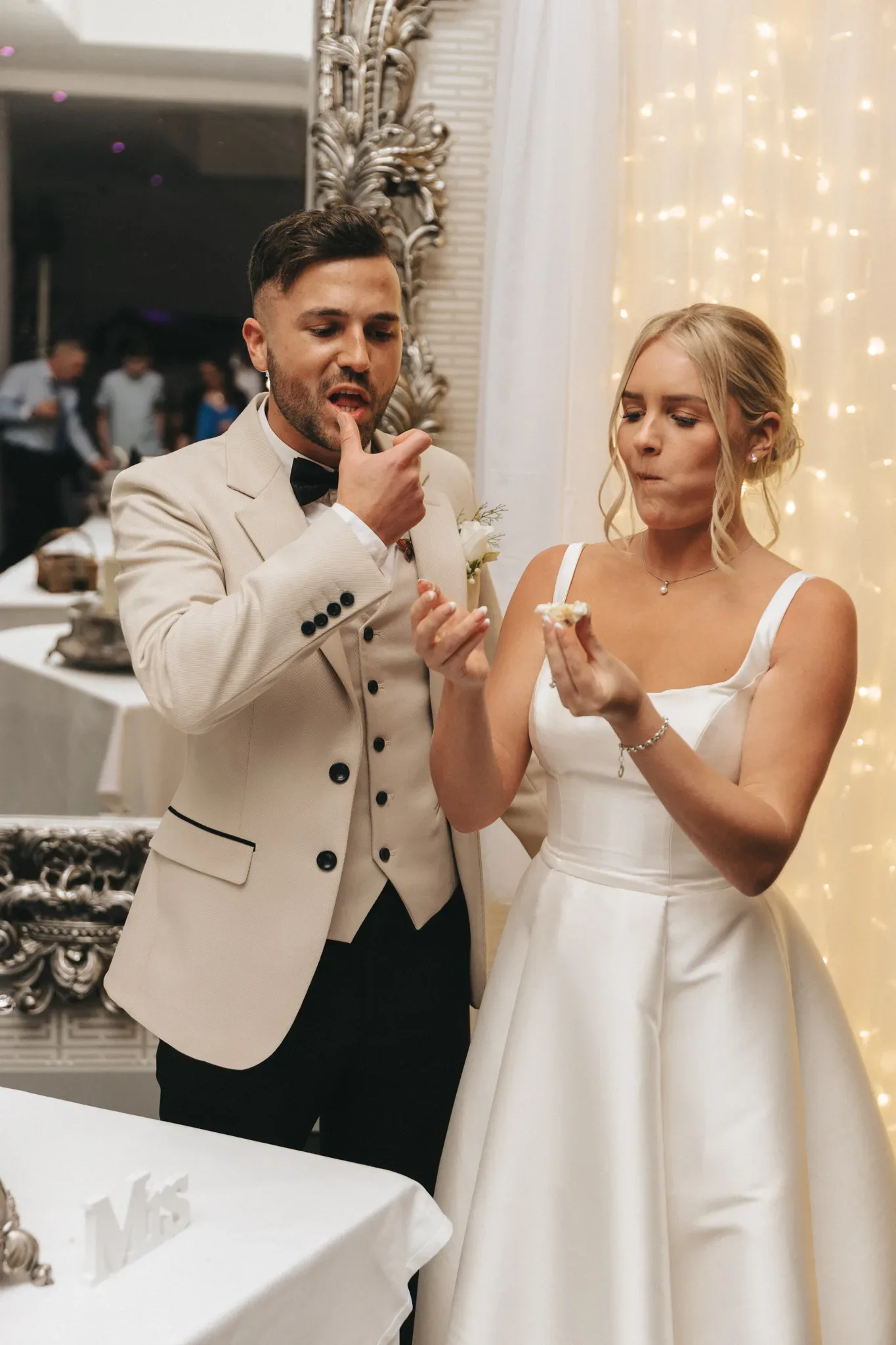 A bride and groom in elegant wedding attire sample food during their reception. the groom, in a cream suit, appears thoughtful, while the bride in a white gown looks unimpressed. a luxurious mirror and fairy lights are in the background.